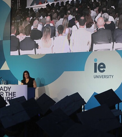 An individual is giving a speech at a graduation ceremony in front of an audience, with IE University branding visible.