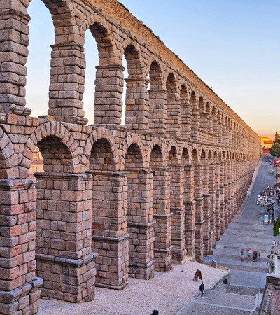 The image depicts a historic aqueduct with tall stone arches in an urban setting during sunset.