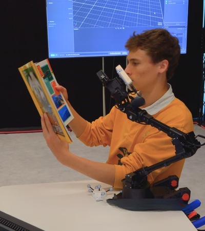A person is using a robotic arm to hold and read books while seated at a desk.