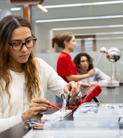 A woman is working on an electronic project at a modern workspace with colleagues in the background.