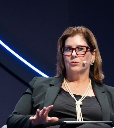 A woman wearing glasses speaks into a microphone while sitting at a conference.