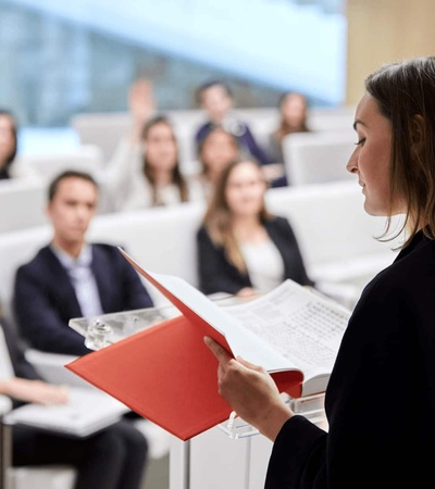 A woman is presenting to an audience in a seminar hall, holding a red folder and addressing attendees who are seated and listening attentively.