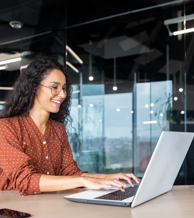 A woman in a polka dot shirt smiling and working on a laptop in a modern office environment.