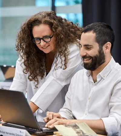 Two professionals, a man and a woman, are smiling and looking at a laptop screen in a modern office setting.