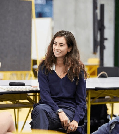 A young woman in a navy blue outfit smiling and sitting on a desk in a classroom setting with peers around.