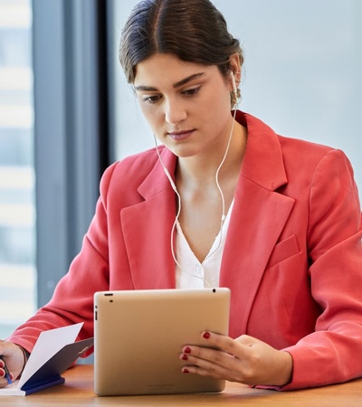 A woman in a pink blazer is working on an iPad while taking notes at a table.