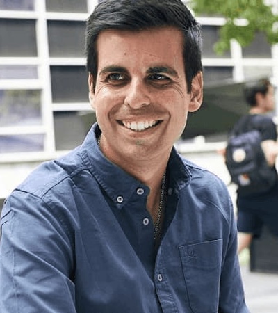 A smiling man in a blue shirt sitting at an outdoor café with modern buildings in the background.
