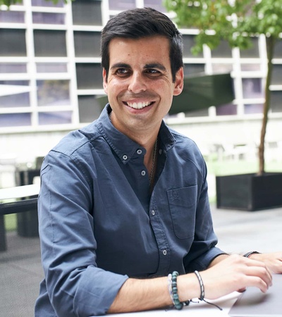 A smiling man working on a laptop at an outdoor table in a modern urban setting.