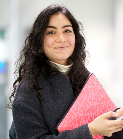 A smiling young woman holding a notebook and a phone in a modern office environment.