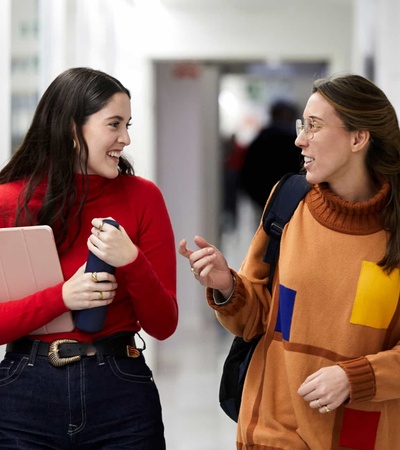 Two young women happily conversing while walking through a school corridor, one holding a notebook and the other a cup.