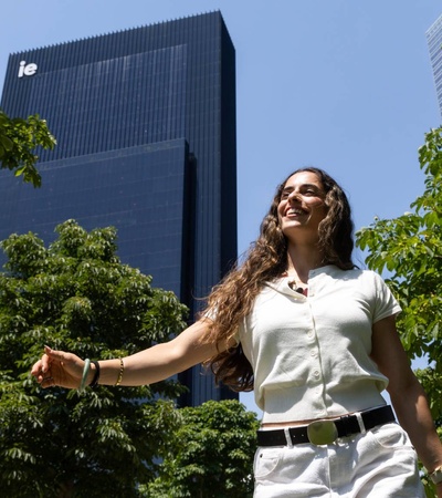 A smiling woman walking among green trees with a modern building in the background.
