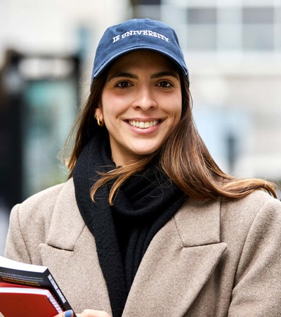 A smiling woman wearing a university cap and a beige coat holds books, standing outdoors.