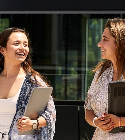 Two women are walking and laughing together outside a modern building.