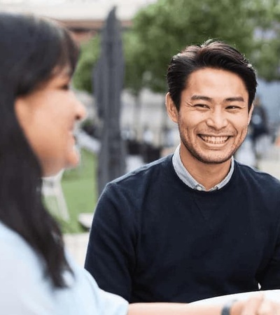 A man smiling at a woman while sitting at a table in an outdoor urban setting.