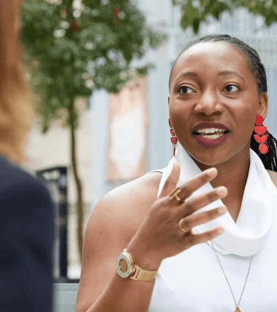 A woman in conversation outdoors, wearing white attire and red earrings, enthusiastically gesturing with her hand.