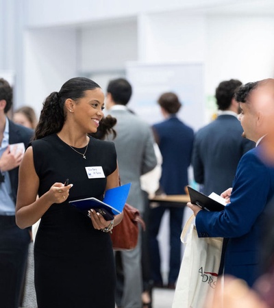 A woman with a name tag smiles while holding a notebook and interacting with others at a networking event.