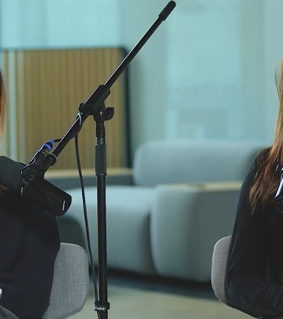 Two women smiling and sitting at a podcast setup with microphones in a modern office space.