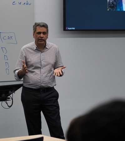 A man is giving a presentation in a classroom with a whiteboard showing diagrams behind him.