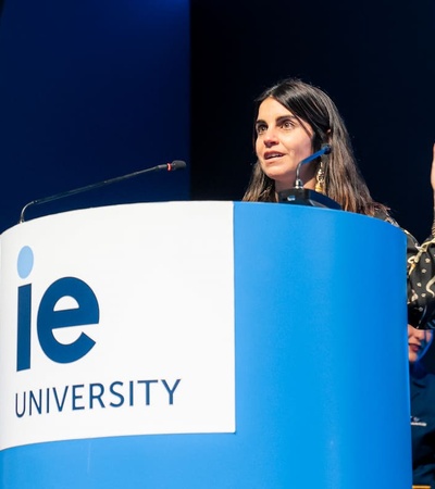 A woman is speaking at a podium with the 'IE University' logo, while listeners in academic attire are present.