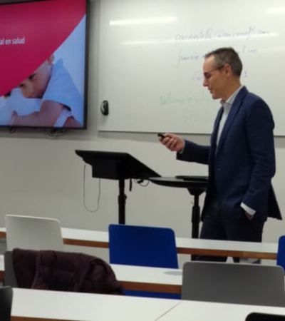 A man in a suit presenting in a classroom with a slide about artificial intelligence in health projected behind him.