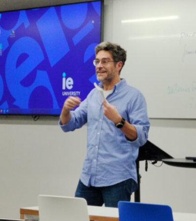 A man is giving a presentation in a classroom with a digital screen displaying the desktop background.