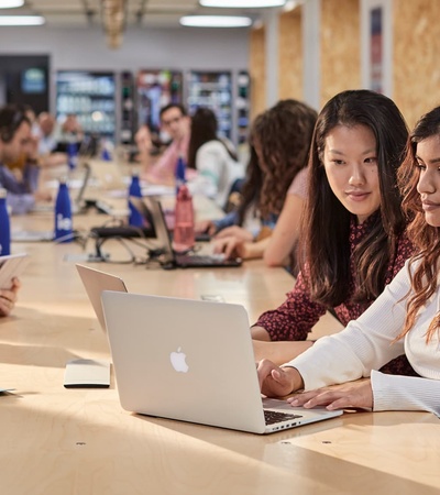 Two women collaborate over a laptop in a spacious co-working environment filled with people.