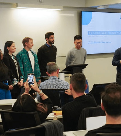 A group of people, including a presenter, stands at the front of a seminar room addressing an audience.