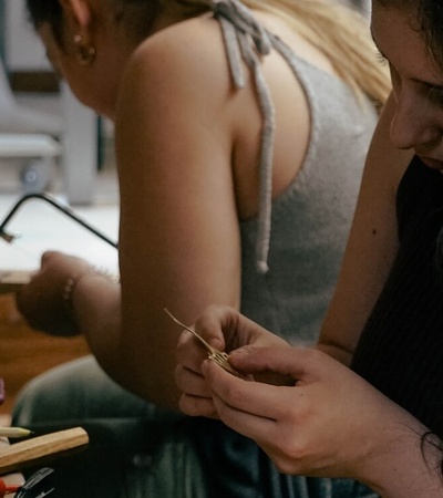 Two women are focused on crafting jewelry at a workshop table cluttered with tools and materials.