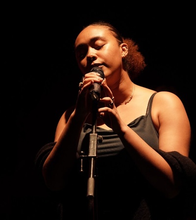 A woman singing into a microphone on a dark stage, illuminated by a soft spotlight.