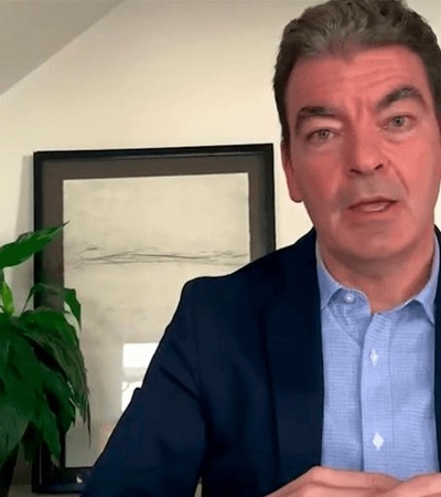 A man in a blue blazer and light blue shirt is speaking, sitting in front of an interior background with a green plant and framed pictures.