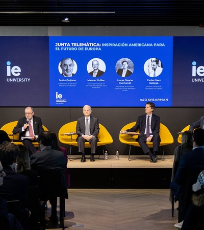 Four men are seated on stage at a panel discussion in a modern conference setting with logos of IE University and monitor displays in the background.