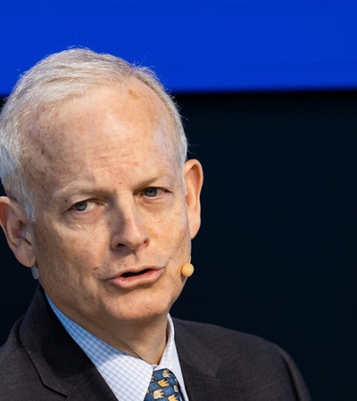 A mature man with gray hair, wearing a suit and tie, is speaking at a podium with a blue and yellow background.