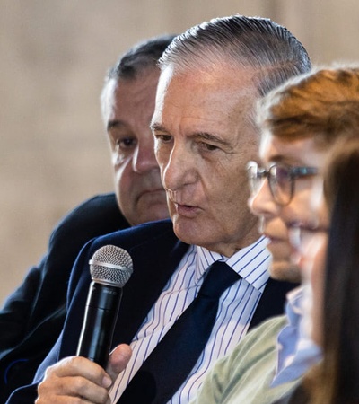 A senior man speaks into a microphone at a panel discussion while seated with other panelists.