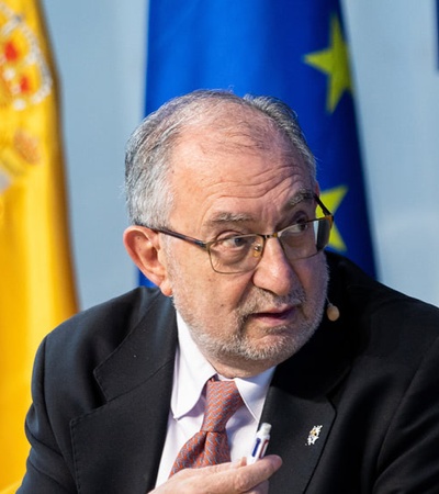 A man in a suit gestures while speaking, with the flags of Spain, the European Union, and Madrid in the background.