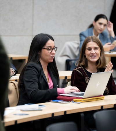 Two women sitting at a desk, using a laptop and interacting in a classroom setting.