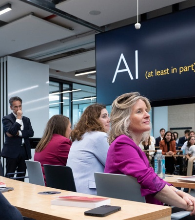 A group of people, likely students and professionals, attentive in a modern classroom setting with an 'AI (at least in part)' presentation on display.