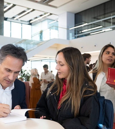 A man signs a book at a table while conversing with a young woman in a crowded event space.