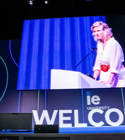 A woman is giving a speech at a podium with a large 'WELCOME' sign on a screen behind her at IE University.