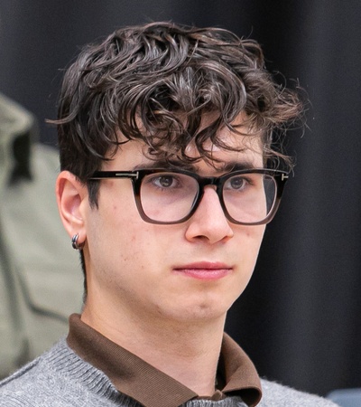 A young man with glasses and curly hair looks thoughtfully to the side.