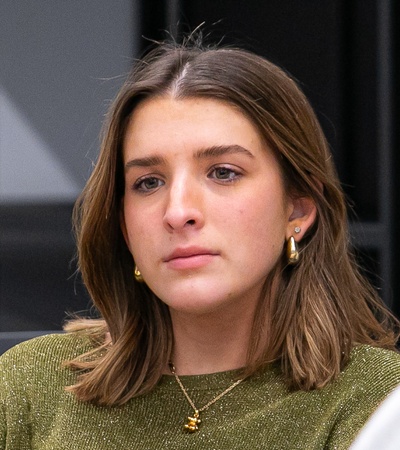 A young woman with brown hair wearing a green top sitting indoors, looking thoughtful.