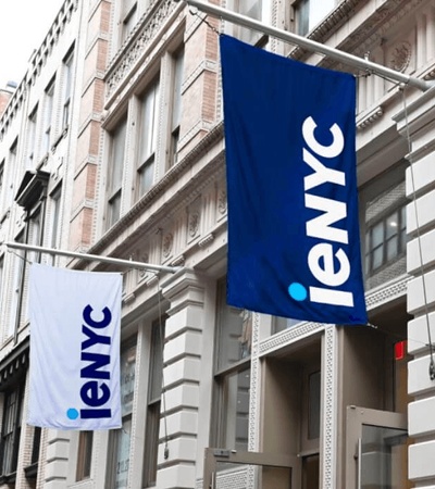 The image shows two flags displaying the logo 'ieNYC' outside a building.