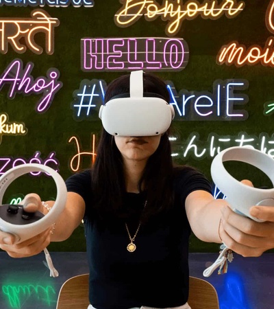 A woman wearing a virtual reality headset stands in front of a colorful wall with 'hello' written in multiple languages.