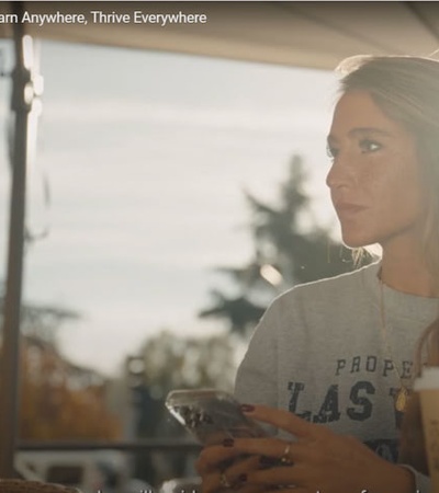 A woman sitting at a cafe table outdoors, looking away while holding a smartphone, with a gentle sunlight illuminating her face.