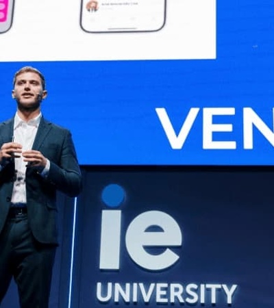 A man in a suit speaking at a podium with the 'IE University VENTURE' banner in the background, with a woman seated beside him.