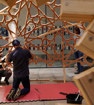 Two people are assembling a large geometric wooden structure outdoors.