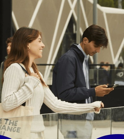 A group of people interacts in an urban setting, with one person smiling at another while others are engaged with their phones.