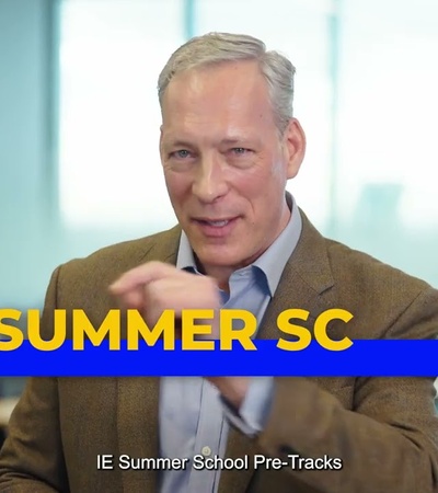 A man is speaking and gesturing in a modern classroom setting, promoting a summer school program.