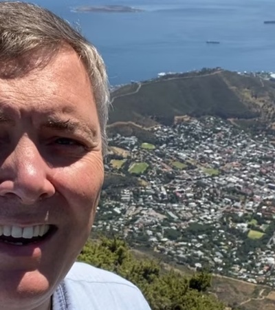 A man takes a selfie with a sprawling cityscape and ocean in the background from a high viewpoint.