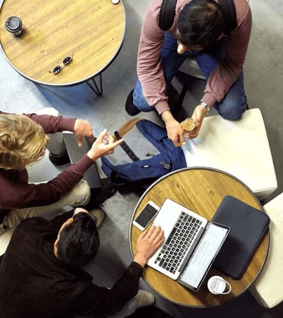 Three people are sitting around a small table in a casual office setting, working together with a laptop and documents.