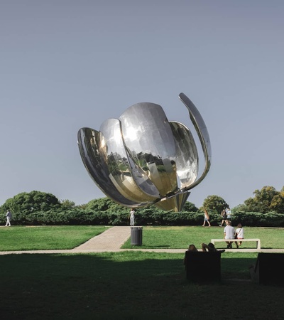 A large metallic sculpture resembling a flower stands at the center of a park, surrounded by people relaxing and enjoying the scenery.